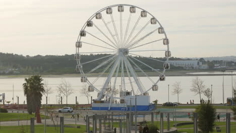 People-Riding-Ferris-Wheel-With-Glass-Cabins-Spinning-In-An-Amusement-Park-By-The-Riverside---wide-shot