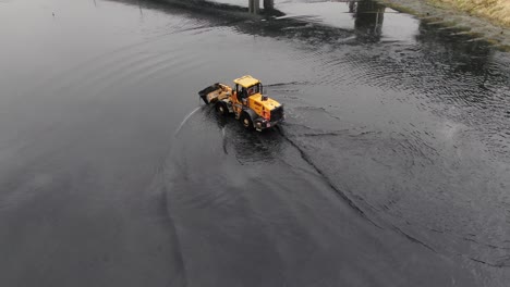 Gelber-Traktor,-Der-Schlamm-Aus-Einem-Kanal-Unter-Einer-Brücke-Schöpft-Und-Reinigt,-Luftbild