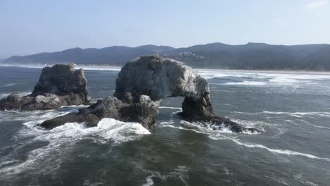 Pulling-away-and-revealing-Twin-Rocks-being-washed-by-rolling-pacific-ocean-waves,-aerial