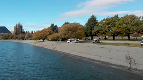 Slowmo-Aerial-Drone-flying-towards-a-campervan-on-a-beach-above-beautiful-blue-lake-in-Lake-Wanaka,-New-Zealand-in-autumn