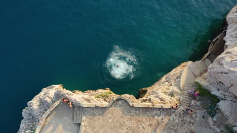 Male-cliff-jumping-backflip-off-rocky-coastal-clifftops,-aerial-top-down-view