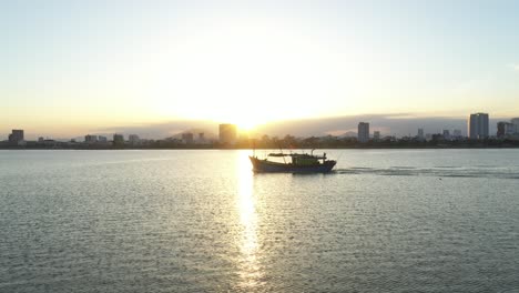 Ein-Fischerboot-Bei-Sonnenuntergang-Mit-Der-Skyline-Von-Vietnam-Im-Hintergrund