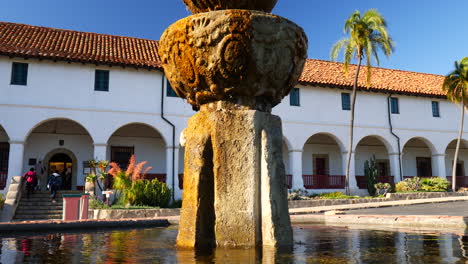 People-and-tourists-walking-up-the-steps-of-the-historic-Santa-Barbara-Mission-building-with-Spanish-Catholic-architecture-and-fountain-in-the-foreground-SLIDE-LEFT