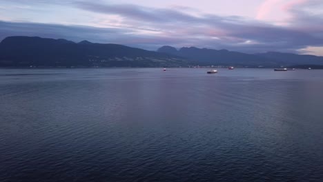 Aerial-Sunset-Wide-Shot-Flying-Over-Pacific-Ocean-Towards-North-Shore-Panning-Right-To-Reveal-Cargo-Ships-And-Downtown-City-Skyline-In-Vancouver-British-Columbia-Canada