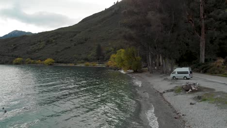 SLOWMO---Motorhome-by-beautiful-blue-Lake-Wakatipu,-Queenstown,-New-Zealand-with-mountains-fresh-snow-cloudy-sky-in-background---Aerial-Drone