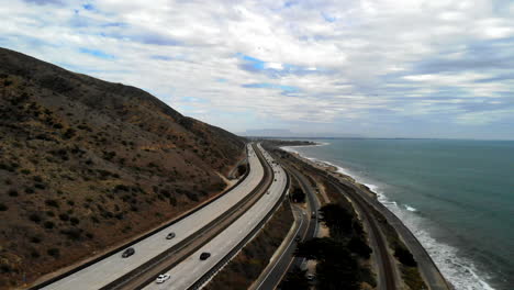Aerial-drone-shot-over-the-101-freeway-with-cars-next-to-the-blue-Pacific-Ocean-waves-and-California-coast-cliffs-in-Ventura