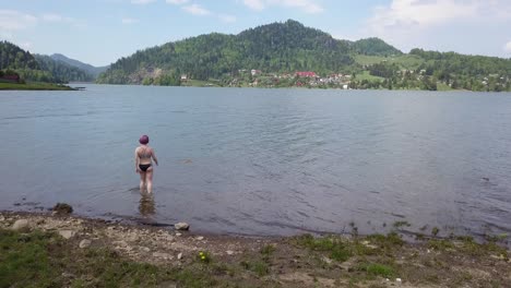 Aerial-approach-shot,-of-girl-with-short-purple-hair-walking-into-lake-with-black-swimsuit-on