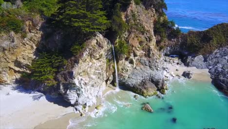 Aerial-drone-shot-over-a-waterfall-cascading-from-rocks-down-onto-the-beach-of-a-remote-cove-with-the-ocean-in-background