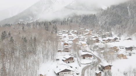 Drone-shot-descending-through-snowy-ski-village,-snow-flying-towards-the-camera-with-low-cloud,-mountains-trees,-and-chalets-in-the-background