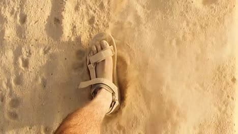 Hombre-Caucásico-De-Primer-Plano-Con-Pies-Polvorientos-Caminando-A-Cámara-Lenta-Con-Sandalias-En-El-Desierto-Arenoso-O-En-La-Playa,-Huellas-Pateando-Algunas-Partículas-Polvo-Brillante-Arena-Usando-Sandalias-En-Un-Día-Soleado