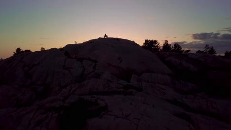 Fly-over-Inukshuk-on-Rocky-Pine-Tree-Island-at-Sunset,-Drone-Aerial-Wide-Dolly-In