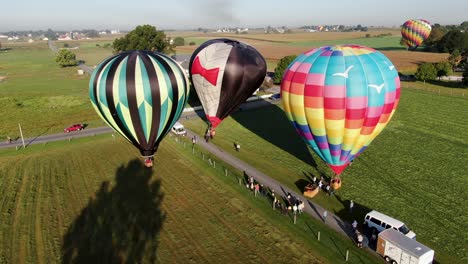 Aerial-turn-reveals-colorful-hot-air-balloons-landing-in-meadow,-passengers-and-onlookers-assist-with-deflating-the-balloons