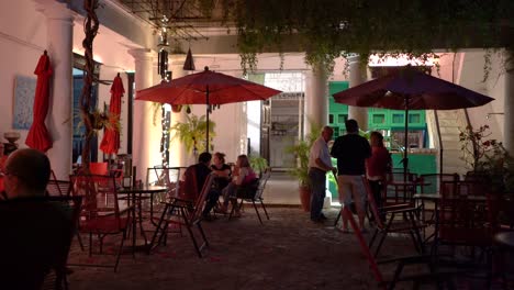 Gente-En-Mesas-Con-Sombrillas-Por-La-Noche-En-Un-Café-Restaurante-Que-Está-Situado-En-El-Patio-Central-De-Un-Gran-Edificio-Antiguo