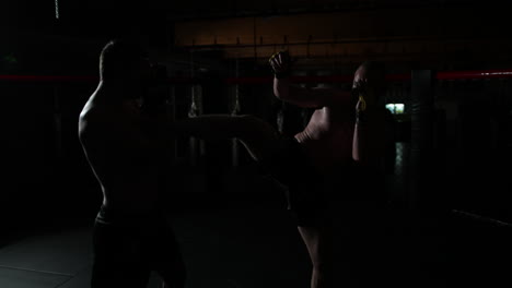 Slow-motion-of-two-mma-fighters,-covered-in-sweat,-fighting-in-a-ring,-one-kicking-the-other-one-and-then-they-immobilize-and-stand-still,-the-camera-goes-around,-in-a-dark-room,-with-a-single-light