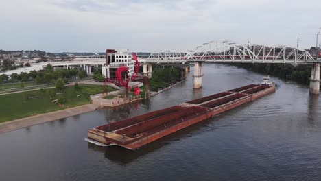 Spectacular-view-of-tugboat-guiding-large-commercial-transport-barge-down-river-beneath-truss-bridge,-Illinois,-aerial