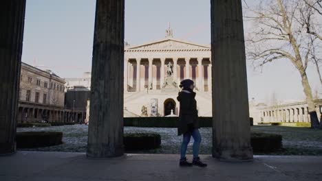 Caucasian-woman-walks-in-front-of-tall-stone-columns-with-imperial-and-impressive-Berlin-UNESCO-Old-National-Gallery-museum-building-in-background,-Germany,-static-profile-slow-motion