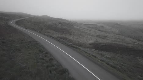 Tomas-De-Drones-De-Patinador-Yendo-Cuesta-Abajo-En-Un-Camino-Sinuoso-En-Un-Paisaje-Brumoso-Y-Dramático-En-Islandia