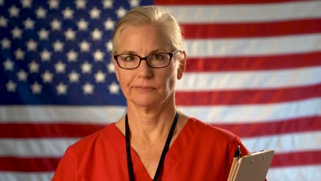 Medium-tight-portrait-of-a-healthcare-nurse-with-clipboard-walking-towards-camera-shaking-her-head-with-an-out-of-focus-American-flag