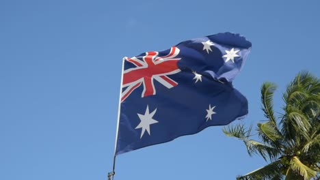 Schöne-Australische-Flagge-In-Zeitlupe-Mit-Wind-Und-Schöner-Palme-Dahinter-Am-Strand-Im-Sommer