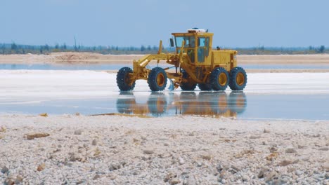 Yellow-Motor-Grader-Driving-And-Working-At-The-Salt-Flats-In-Kralendijk,-Bonaire-On-A-Sunny-Day