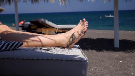 Nahaufnahme-Der-Füße-Einer-Frau-Beim-Sonnenbaden-Am-Lavasand-In-Perissa-Santorini,-Mit-Sonnenschirmen-Und-Dem-Meer-Im-Hintergrund