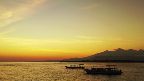 Paradise-tropical-seaside-at-sunset-with-yellow-orange-colors-of-sky-reflecting-on-vibrant-sea-surface,-silhouette-of-anchored-boats-on-bay,-Indonesia