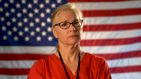 Medium-tight-portrait-of-a-healthcare-nurse-looking-concerned-and-sympathetic-walking-towards-the-camera-with-an-out-of-focus-American-flag