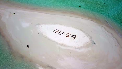People-form-NUSA-letters-meaning-island-in-the-Indonesian-language-on-a-tiny-sandy-isle-in-Raja-Ampat,-Aerial-dolly-out-shot