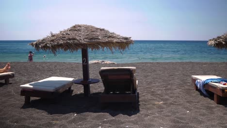 Handheld-Wide-Shot-of-a-Tiki-Hut-By-The-Black-Lava-Beach-in-Perissa-Santorini-Greece-With-Tourists-Swimming-and-Enjoying-The-Beach