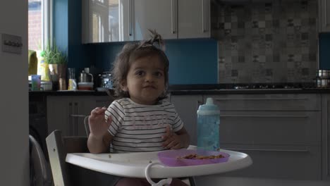 Britisches-Asiatisches-Baby,-Das-Lebensmittel-Von-Einer-Plastikplatte-Auf-Einem-Kinderstuhl-Isst