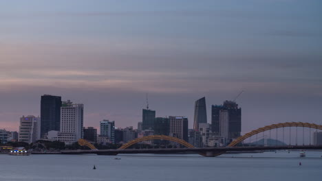 Stadtzeitraffer-Der-Modernen-Städtischen-Skyline---Ein-Übergang-Von-Tag-Zu-Nacht-Mit-Neonlichtern,-Drachenbrücke-Und-Booten-Auf-Dem-Fluss-In-Vietnam,-Asien