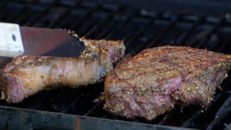 Eine-Fleischzange-Dreht-Ein-Fast-Gares-Saftiges-Rib-Eye-Steak-Auf-Einem-Grill-Und-Lässt-Es-In-Zeitlupe-Ein-Wenig-Gleiten
