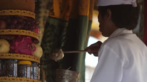 Ofrendas-De-Comida-En-El-Altar-Del-Templo-Hindú-En-Bali,-Indonesia,-Mientras-Un-Sacerdote-Vestido-De-Blanco-Saca-Agua-En-Una-Taza-De-Plata-Para-Bendición-Y-Ceremonia