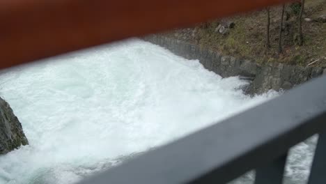 Powerful-white-foamy-water-stream-view-trough-bridge-railing,-Pan-left