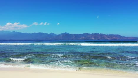 Peaceful-seascape-with-white-waves-of-sea-covering-rocky-seabed-near-white-sandy-beach-on-a-bright-blue-sky-over-mountains-horizon-in-Bali
