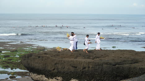 Balinesische-Frauen-Legen-Ihr-Tägliches-Opfer-Dar,-Während-Surfer-Im-Hintergrund-Auf-Wellen-Warten