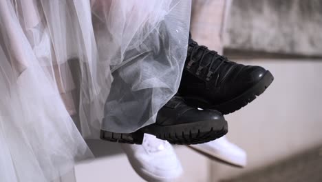Close-up-of-wedding-couple-in-black-boots-and-white-sneaker-swinging-them-while-sitting-on-the-edge-of-stone-wall-warehouse-gate