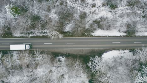 Tracking-a-lorry-from-above-which-is-driving-along-a-straight-wintry-road,-framed-with-trees-from-a-conifer-forest