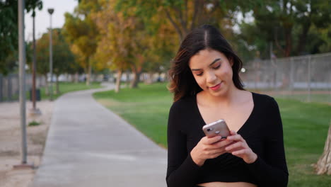 Beautiful-young-independent-hispanic-woman-looking-happy-as-she-checks-her-phone-for-text-messages-or-social-media-notifications-on-a-walk-through-the-park