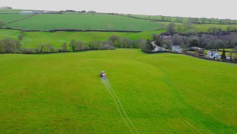 Smooth-aerial-drone-view-following-a-red-tractor-fertilising-a-greet-field-with-trees-from-a-distance