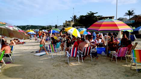 Dolly-in-of-families-sitting-in-the-sand-under-umbrellas-near-the-shore-in-Bombas-and-Bombinhas-beaches,-Brazil