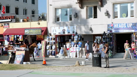 LAPD-on-Horseback-Walking-Down-Venice-California-Boardwalk-on-Sunny-Day,-Pedestrians-Walking-and-Hanging-Out-Around-Storefronts