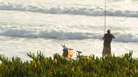 Man-in-Hat-Casts-Fishing-Line-into-the-Ocean-as-Waves-Crash,-Plants-in-Foreground