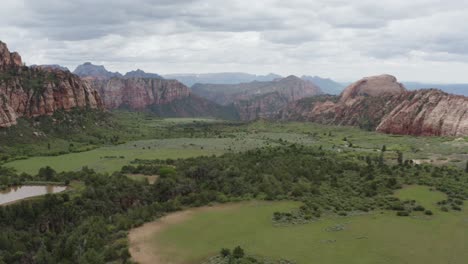 Wide-slow-panoramic-drone-shot-of-Zion-Nation-Park-valley,-green-grass-and-prominent-red-rocks