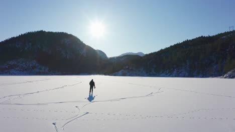 Aerial-following-man-silhouette-in-bright-sun,-ice-skating-on-frozen-lake-Mountains-Background