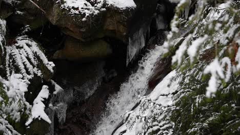 Water-splashing-down-over-a-small-waterfall-between-gray-rocks-while-snowflakes-whirling-down-and-covering-the-nature-into-a-white-snow-winter-scene