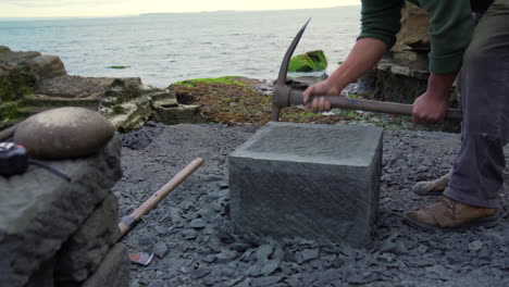 Handheld-wide-shot-of-a-craftsman-shaping-a-square-block-go-cancagua-stone-with-a-stone-pick,-on-the-shoreline-of-the-city-Ancud,-Chiloe-Island