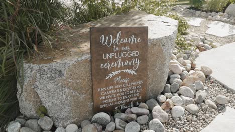 Welcome-to-a-unplugged-ceremony-wood-sign-resting-against-a-large-rock-in-the-middle-of-a-outdoor-garden-ceremony-venue-at-Le-Belvédère-in-Wakefield,-Quebec,-Canada