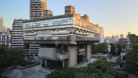 Aerial-dolly-in-of-modern-exposed-concrete-National-Library-surrounded-by-buildings-and-trees-at-sunset,-Buenos-Aires