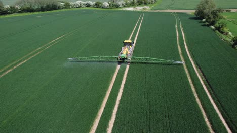 A-drone-shot-following-and-descending-towards-a-yellow-and-green-tractor,-spraying-wheat-fields-with-his-machinery-behind